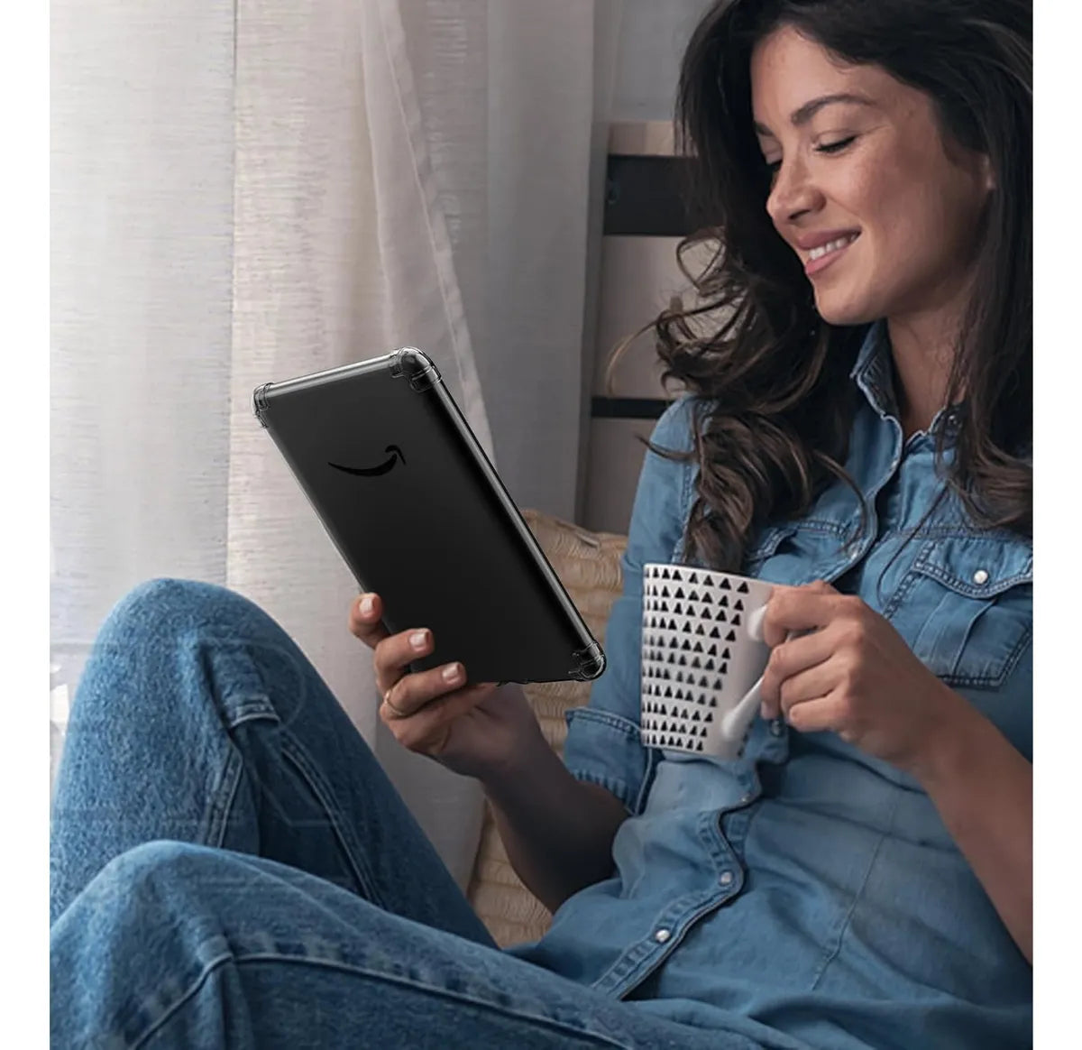 Una mujer vestida de mezclilla está sentada junto a la ventana, sonriendo mientras sostiene una DIETEC Funda Transparente + Mica para Kindle 2024 11va Gen 6” en una mano y una taza estampada en la otra.